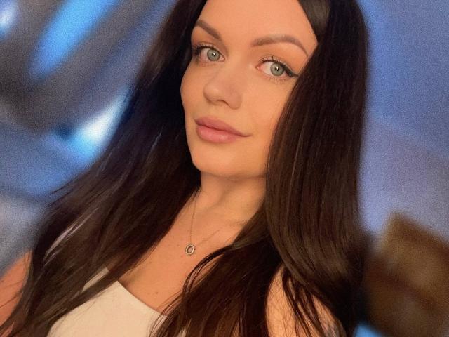 AnastasiaMorante with long, dark brown hair, green eyes, and pink lips, wearing a white top, has a small necklace, and a subtle smile, in a blurry background.