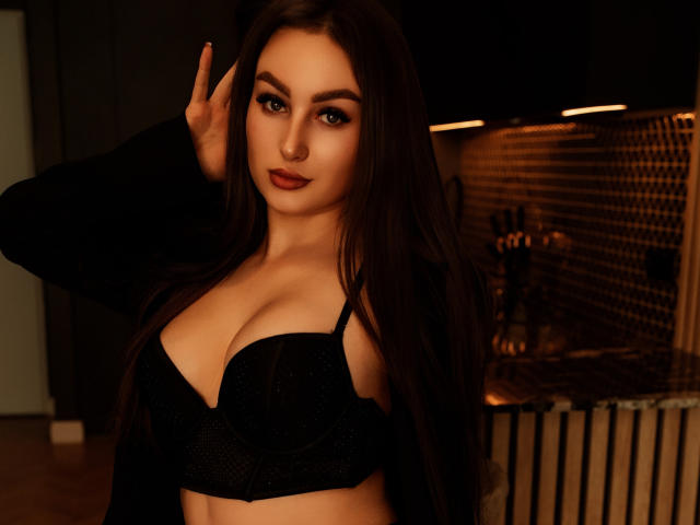 DonnaBeauty with long brown hair, dark eyes, and red lipstick, wearing a black bra that highlights her big, round tits, and a black jacket, making a peace sign.