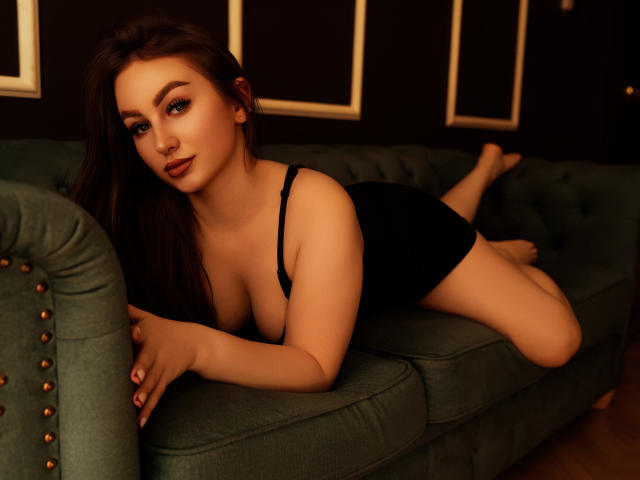 DonnaBeauty with long brown hair, lying on a green velvet couch, wearing a black dress, showing her tits, with a seductive smile, and her ass slightly raised.