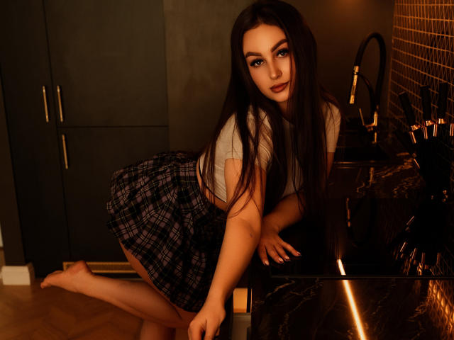 DonnaBeauty with long dark hair, blue eyes, and a cute smile, wearing a grey top and black plaid skirt, leaning on a dark kitchen counter, showing her ass.