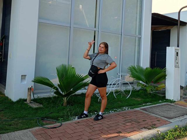SamantaGreen standing outside, holding a broom, wearing a gray tee, black shorts, black sneakers, and a black crossbody bag, with a white bicycle and green plants behind her.
