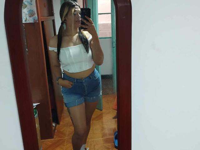 SamantaGreen in a mirror selfie, wearing a white off-shoulder top, denim mini-skirt, and white socks, with long black pigtails, holding a phone, showing her tits and ass.
