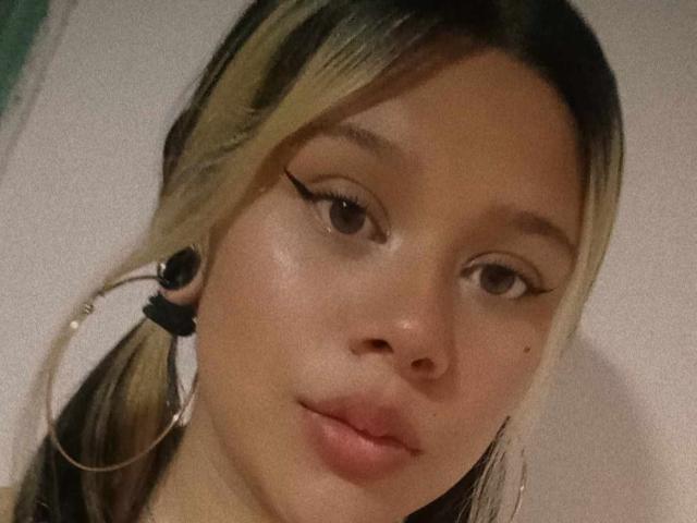 SamantaGreen's face with blonde and black hair in pigtails, wearing large hoop earrings, has defined eyebrows, eyeliner, and slightly parted pink lips, with a small mole on her right cheek.