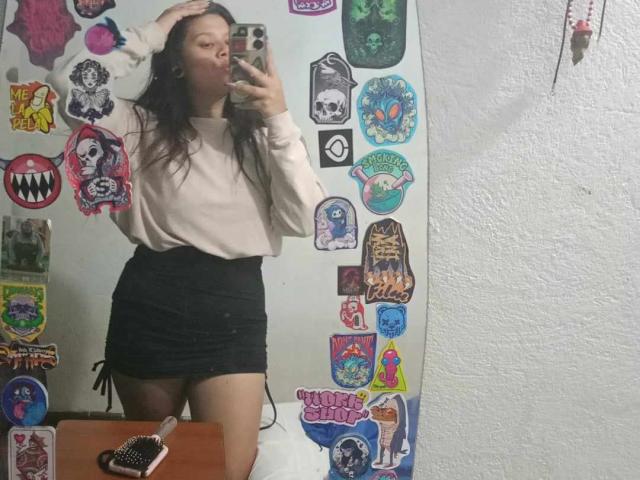SamantaGreen with long brown hair, white sweater, black skirt, taking mirror selfie, hand on head, showing off her ass, stickers on wall, phone in hand, mirror reflection.