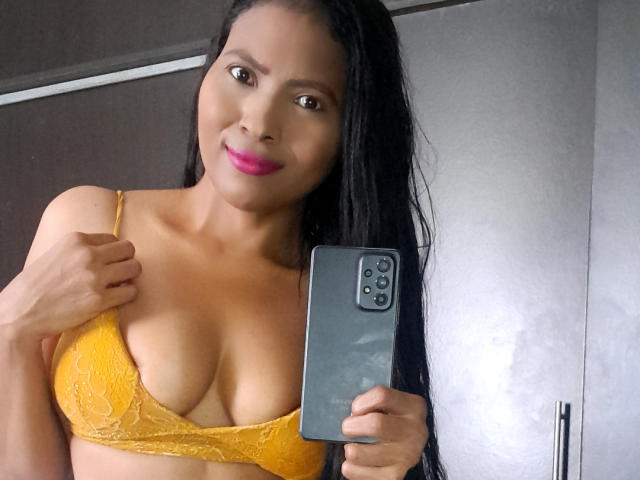 DulceMiel69 with long black hair, yellow lace bra, pink lipstick, holding a smartphone, showing her boobs, smiling, tan skin, in a mirror selfie, indoors, holding bra strap.