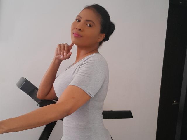 DulceMiel69 with brown skin, black hair in a bun, wearing a grey t-shirt, smiling, touching her chin, standing on a black exercise bike, looking at the camera.