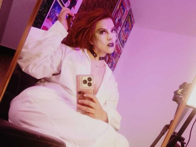 SkylarDesire with red hair, wearing a white robe, purple lipstick, and a choker, taking a mirror selfie with a pink phone, sitting on a black chair.
