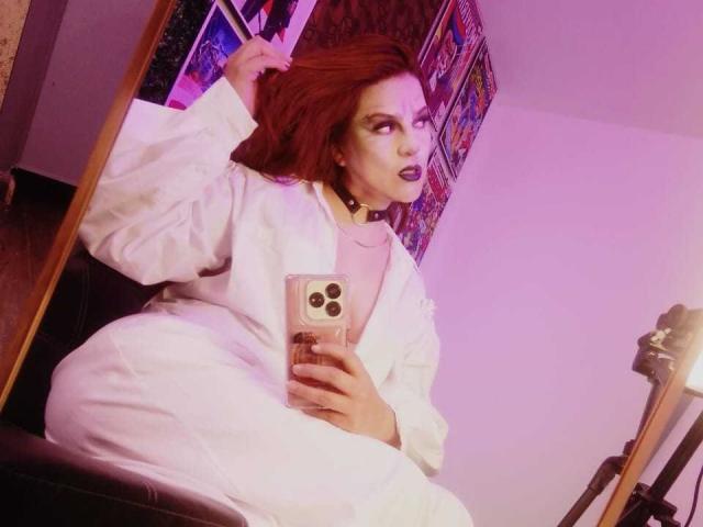 SkylarDesire with red hair, wearing a white robe, choker, and pink top, holding a phone, sitting in front of a mirror, looking at herself with a seductive expression.
