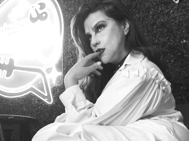 SkylarDesire with long hair, wearing a white shirt, biting her finger, looking seductively at the camera in a black-and-white photo. Her tits are subtly visible under the shirt.