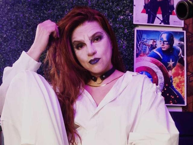 SkylarDesire has red hair, wearing a white shirt with puffy sleeves, black choker, and grey lipstick, touching her hair with a serious expression, posters of superheroes behind her.