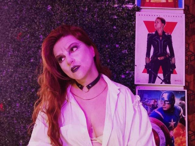 SkylarDesire with red hair, black choker, white shirt, and purple lipstick, standing next to Marvel posters of Jessica Jones and Captain America. Boobs slightly visible under shirt.