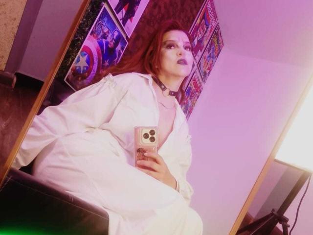 SkylarDesire with red hair, dark lipstick, wearing a white robe, holding a phone, sitting on a black leather seat, pink and purple lighting, mirror reflection. Boobs not visible.