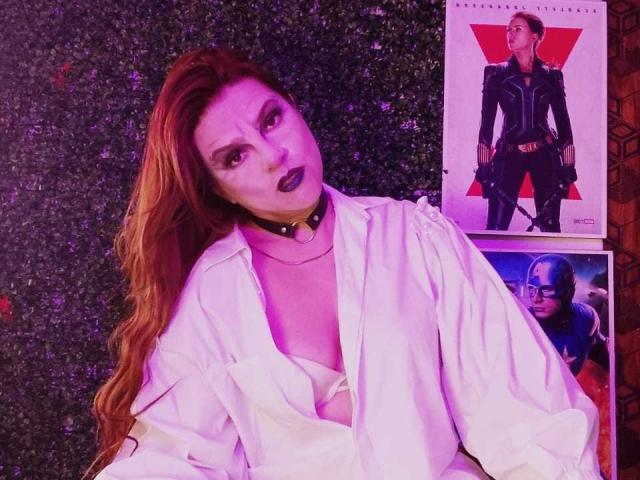 SkylarDesire with red hair, black choker, white shirt, and open chest, looking up, surrounded by movie posters, one showing her in a black suit. Purple lighting.