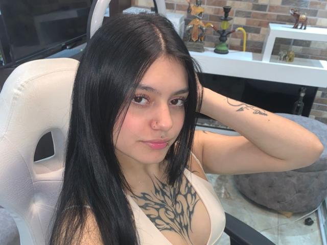 KristyAss with long black hair, nose ring, and tattoo on chest, sitting in white chair, looking at camera, showing large tits, wearing white top, hand in hair.