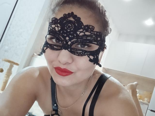 AlinasissyX with black lace mask, red lipstick, and black strap lingerie, showing her tits and smiling, in a bright room. Her brown hair is up, and she's wearing a necklace.