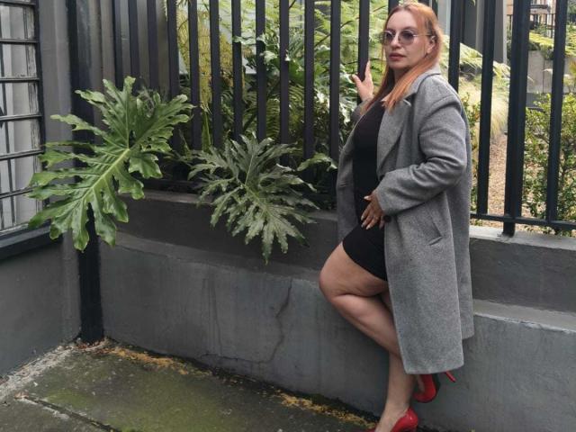 RoxieJackson stands against a black fence, wearing a grey coat, black dress, red heels, and glasses, with green plants beside her, her ass slightly raised, showing her tits.