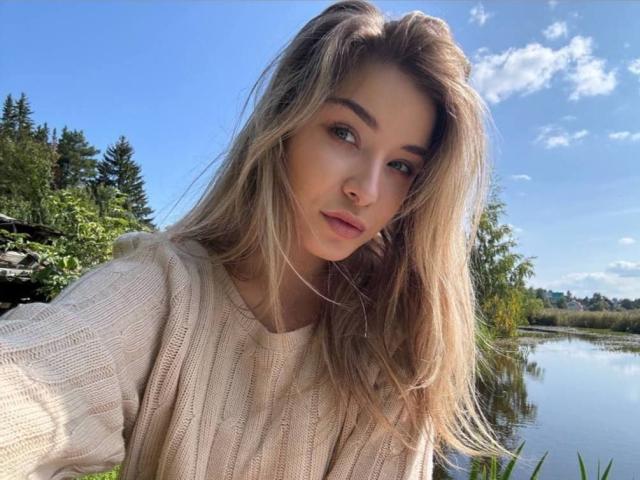 Ariemel with blonde hair, green eyes, and light makeup, wearing a beige knit sweater, standing by a reflective river under a blue sky with scattered clouds.