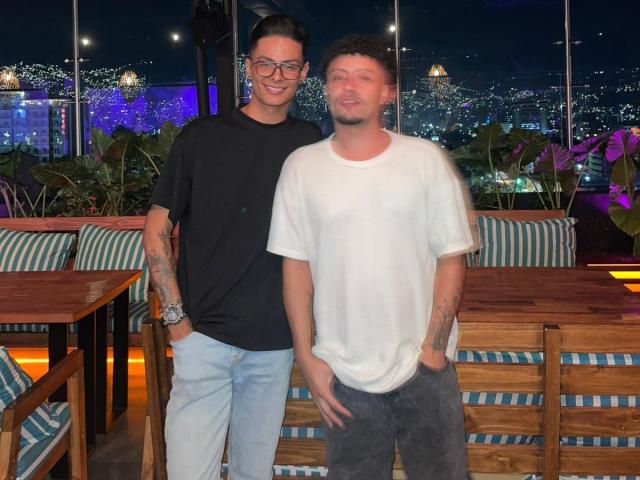 Dos hombres de pie, uno con tatuajes, gafas, camisa negra y jeans azules, el otro con camisa blanca y pantalones negros, ambos con las manos en los bolsillos, sonriendo por la noche..