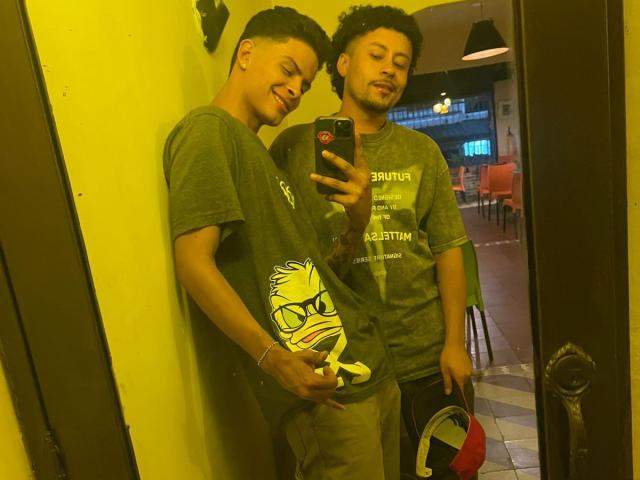 Dos hombres de pie en una habitación iluminada con luz amarilla, uno tomando una foto con el móvil, vistiendo camisetas verdes, con una mano en el trasero del otro, sosteniendo una bolsa con un emblema rojo..