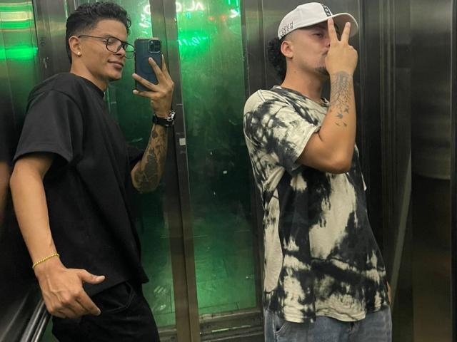 Dos hombres en un ascensor. Uno con camisa negra, gafas y pulsera, sosteniendo un teléfono. El otro con una camisa de colores, una gorra blanca y haciendo el gesto de la paz. Ambos tienen tatuajes..