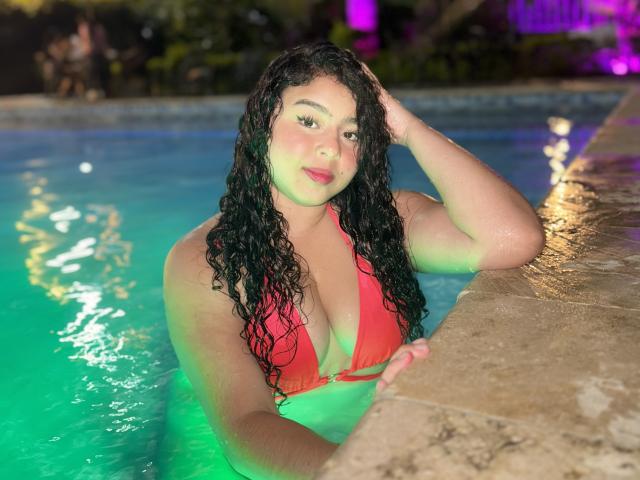 AprielKeith with wet curly black hair, red bikini, large boobs, and green-blue lit pool, leaning on stone edge, hand in hair, smiling at camera.