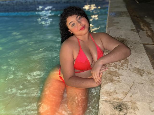 AprielKeith, curvy with large tits in a red bikini, has tan skin and curly black hair, leaning on a pool edge, water up to her thighs, sexy and confident.