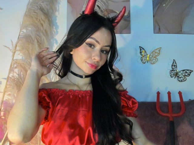 KattRouse with long black hair, red devil horns, red off-shoulder dress, black choker, and pink lipstick, holding hair, standing, with yellow butterfly wall decorations and red trident.