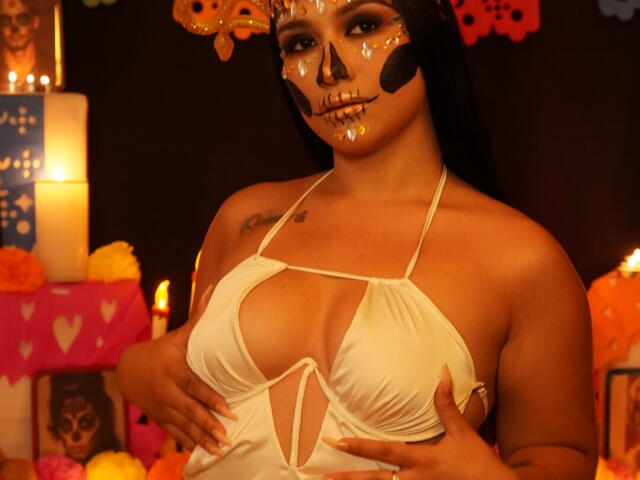 CamilaVerone with painted skull face, white halter top showing large tits, hands on her chest, black hair, orange skull headpiece, candles, flowers, and decorations in the background.