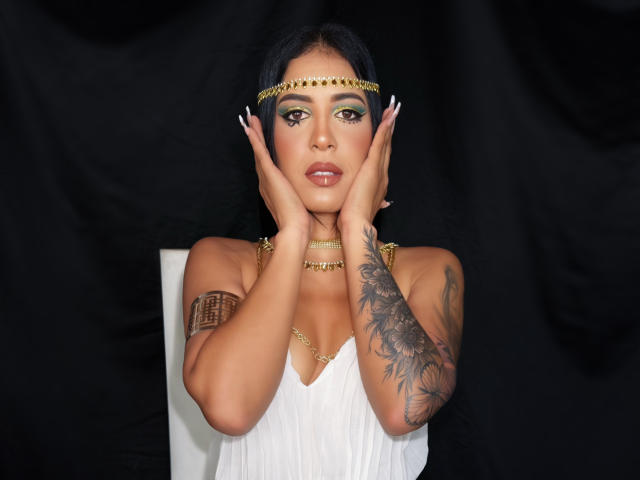 JoannaCool with black hair, gold headpiece, and white dress, hands on face, gold necklace, arm tattoo, and long nails, against black backdrop, showing sexy brown eyes and small tits.