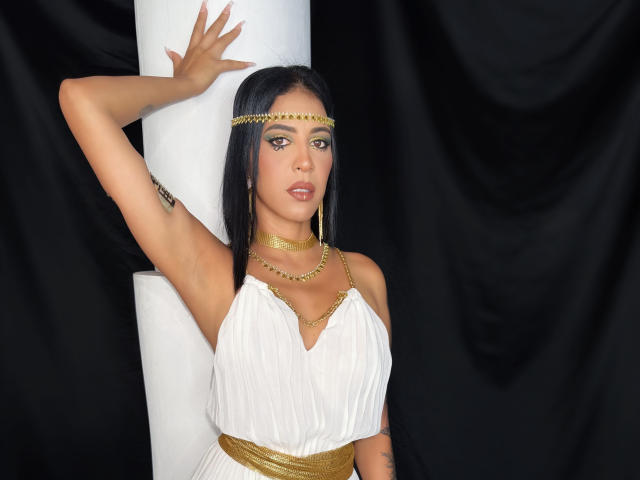JoannaCool with long black hair, gold headband, and white dress, arm raised, showing her small tits and gold necklace, black background, white pillar, and gold waistband.