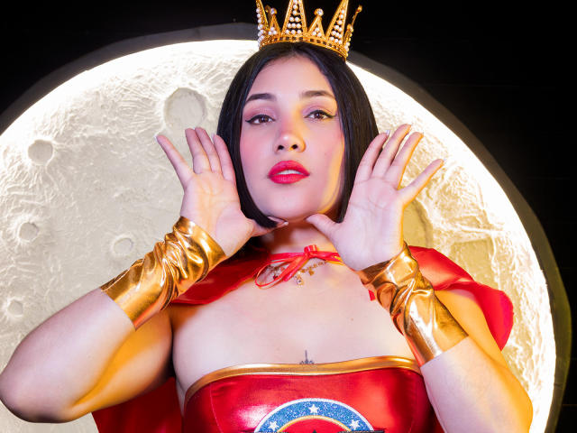 SamantaMuller in a red and gold superhero costume with a crown, hands up, pink lipstick, black bob, gold bracelets, moon background, and a small tattoo on her chest.