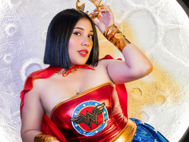 Samanta Muller vestida con un brillante traje rojo de Wonder Woman, con brazaletes dorados y falda azul, sosteniendo una tiara dorada, tiene un corte de pelo bob negro, labios rojos y pechos visibles..