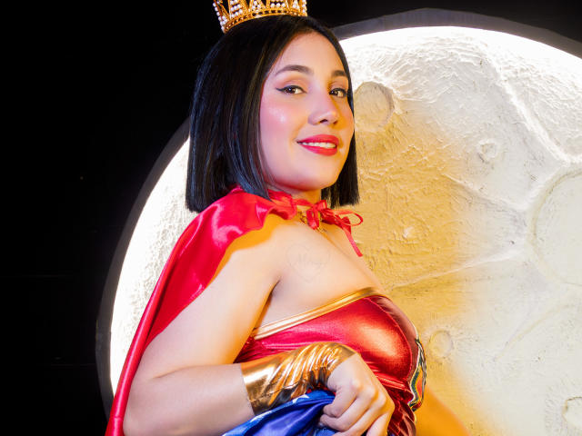 SamantaMuller with a cute smile, wearing a Wonder Woman costume, red cape, gold crown, and shiny red top, holding blue skirt, showing her tits, with black bobbed hair.