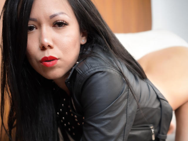 KateOwenns with black hair, red lips, and black leather jacket, leaning forward, showing her bare ass. She's looking at the camera with a seductive expression.