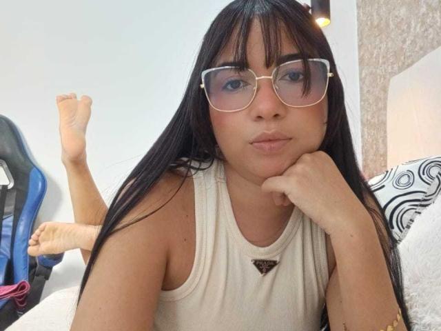 AliceSmity with long black hair, wearing glasses, white tank top, pouty lips, and resting chin on hand, legs up, in a chair, bare feet.