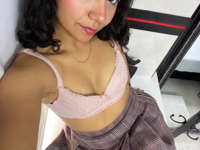 JuliaBennett with medium brown skin, curly black hair, pink lace bra showing small tits, pink plaid skirt, and pink lipstick, wearing a gold chain, taking a selfie.