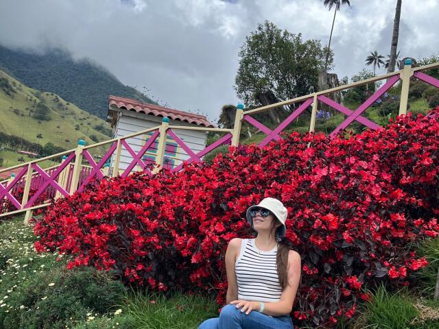 AnnaFer with red bush tits, wearing a white bucket hat and striped tank top, sitting in front of a pink and yellow wooden fence, looking up.