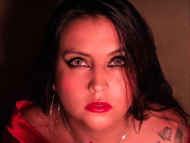 MissSusan has red lips, dark eyes with heavy eyeliner, and black wavy hair. She's wearing a red top and silver hoop earrings, with a tattoo on her right shoulder.