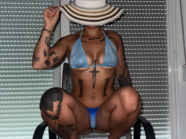 Daphnenights-hot in a blue bikini, squatting with legs apart, wearing a wide-brimmed hat, showing multiple tattoos, including a sword on her chest and a rose on her thigh.