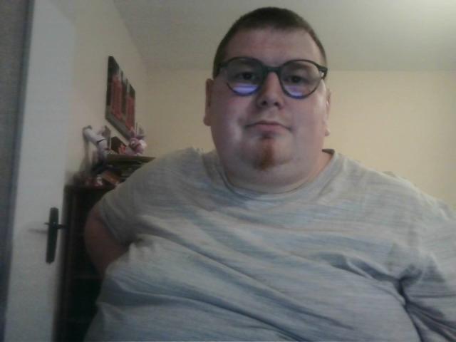 A chubby, bald, bespectacled LoveDoux with a goatee wearing a grey shirt, slightly squished, standing in a dimly lit room.