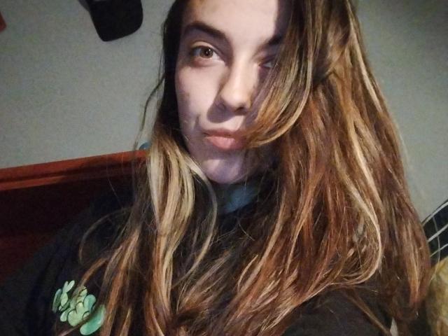 LaMorena69 with long, wavy brown hair, slight smile, and brown eyes, wearing a black shirt with a green design, in a dimly lit room, looking at the camera.