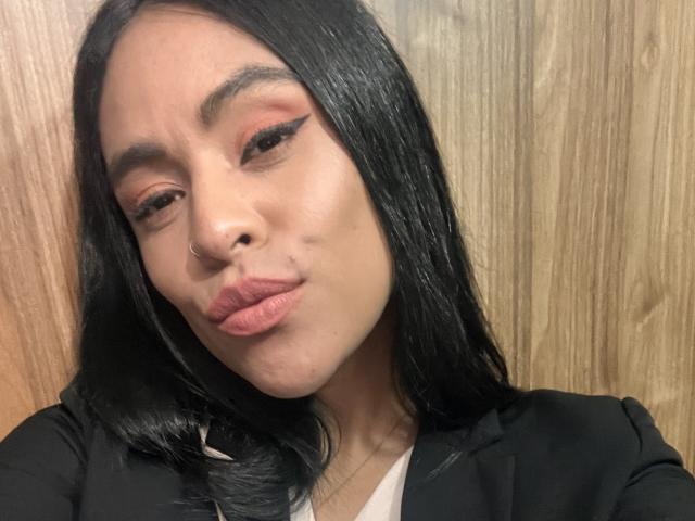 StellaBeckker has black hair, a nose ring, and thick eyeliner. She's pouting her lips with pink lipstick. She's wearing a black top. Wooden background.