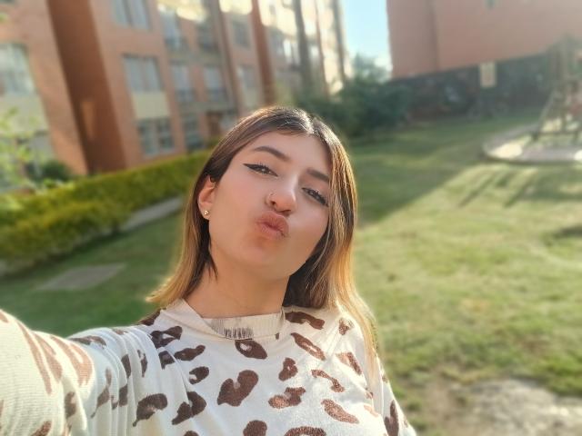 Kaiix a les lèvres pulpeuses, des cheveux bruns clairs, et porte un pull blanc avec des taches de léopard brunes. Elle est à l'extérieur, avec la lumière du soleil sur son visage, et elle a l'air adorable..