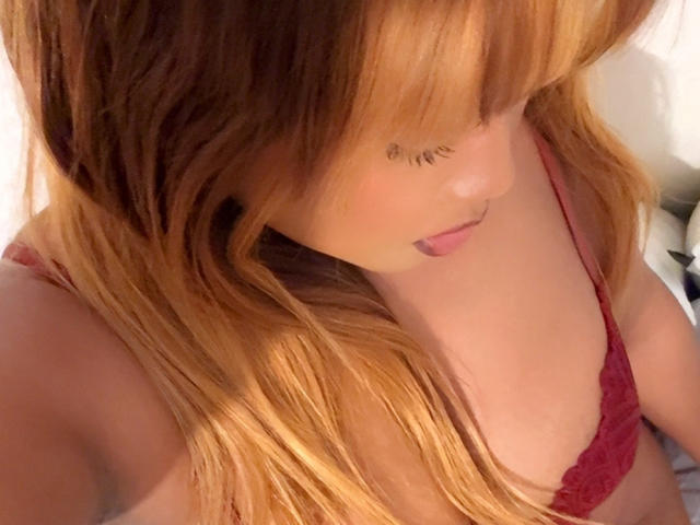 Elmayala, avec ses longs cheveux ondulés et roux, regarde vers le bas, portant un soutien-gorge en dentelle rouge, laissant apparaître ses épaules nues et une partie de sa poitrine. Ses lèvres sont légèrement entrouvertes..
