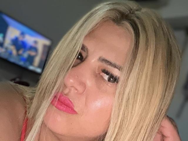 Blonde Raluq with pink lips, heavy black eyeliner, and long lashes, making a pouty face, wearing a red top, her hand in her hair, looking into the camera.