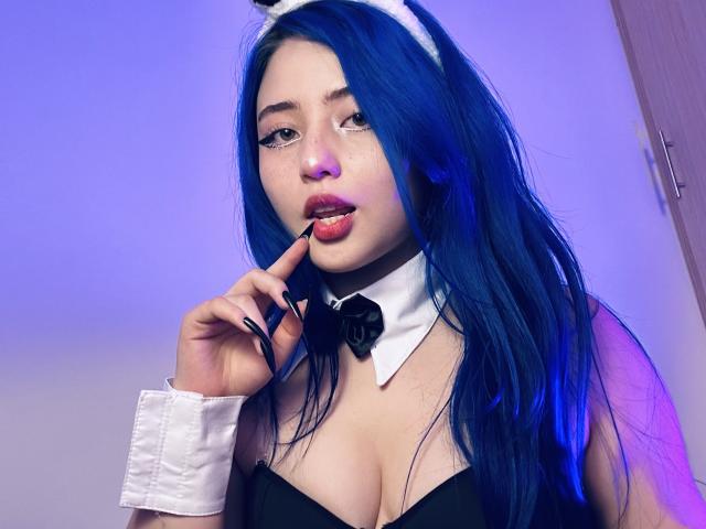 AmeliaXWalker with blue hair, white bunny ears, black strapless top, white collar, black bowtie, and wrist cuffs, sucking on a lipstick applicator, showing her pink lips and cleavage.
