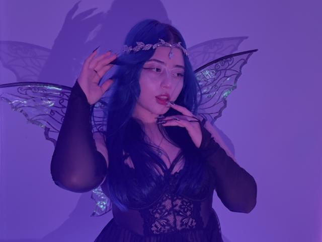 AmeliaXWalker with blue hair, fairy wings, and a jeweled headpiece, touching her lips, wearing a black lace dress, in purple lighting, showing her tits and fingers.