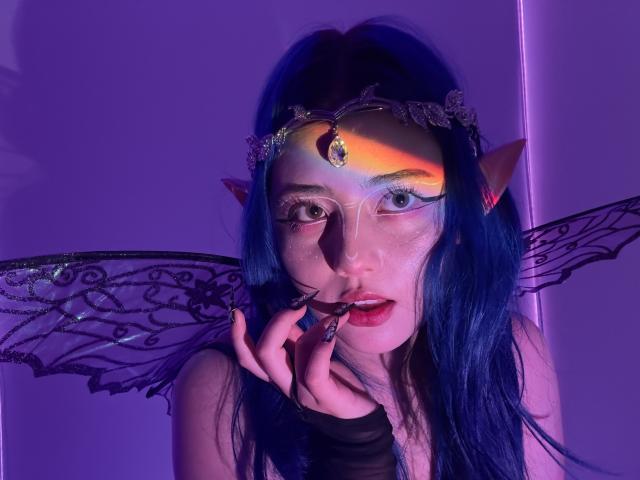 AmeliaXWalker with blue hair, elf ears, and fairy wings, has a purple and orange light on her face, biting her finger with red lips, wearing a delicate headpiece.