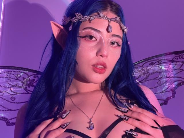 AmeliaXWalker with blue hair, elf ears, fairy wings, glitter on face, jeweled headpiece, and black lingerie, holding her tits with black-nail, glitter, and jeweled fingers.