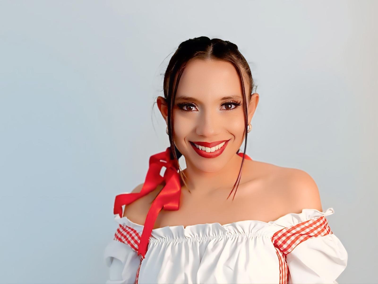 PerlaVonRose with brown eyes, red lipstick, and white off-shoulder top with red ribbons, smiling at the camera, red ribbon in hair, and bare shoulders, looking cute.
